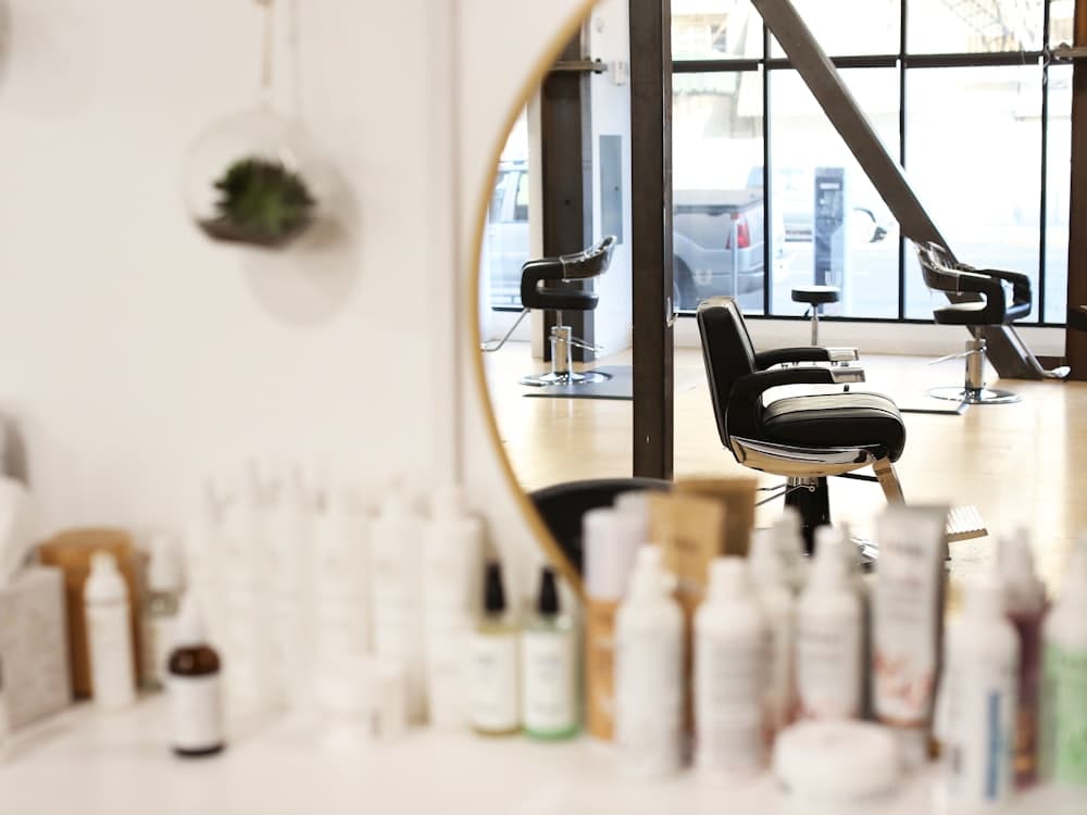 A bright hair salon with a round mirror, styling chair and shelves of products.