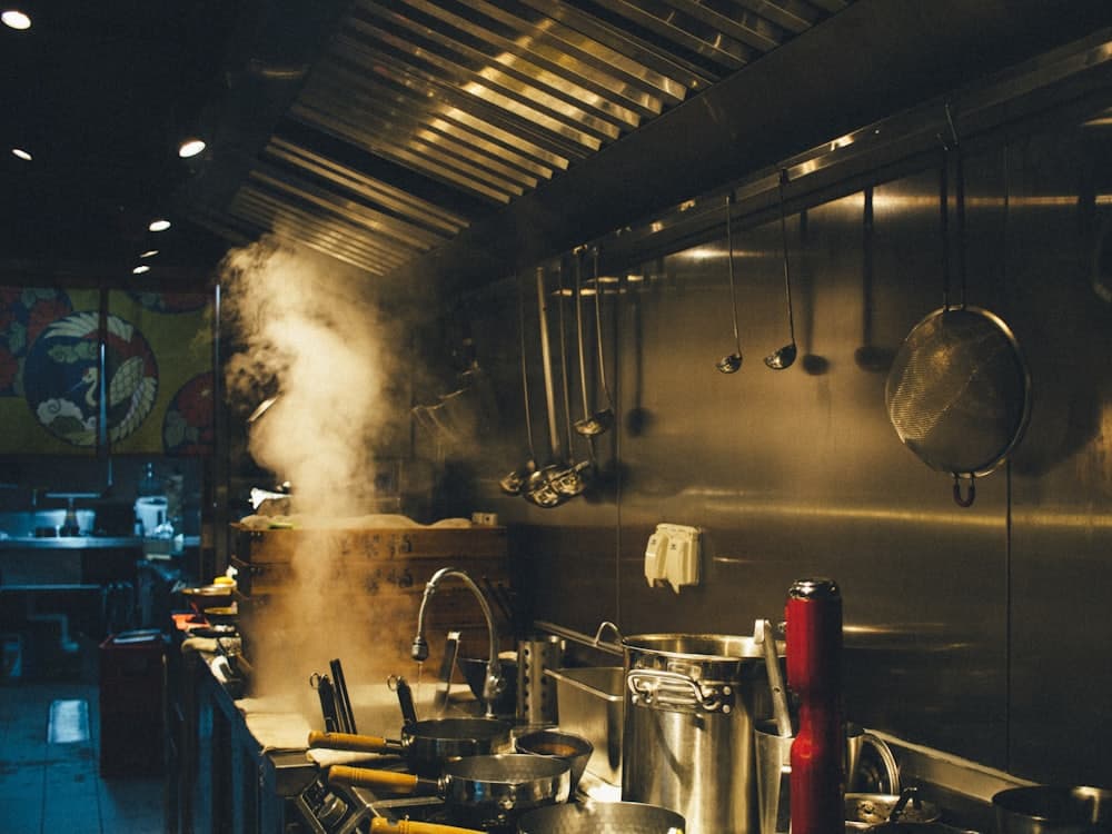 A busy commercial kitchen with steam rising from a pan and pots stacked along the counter.